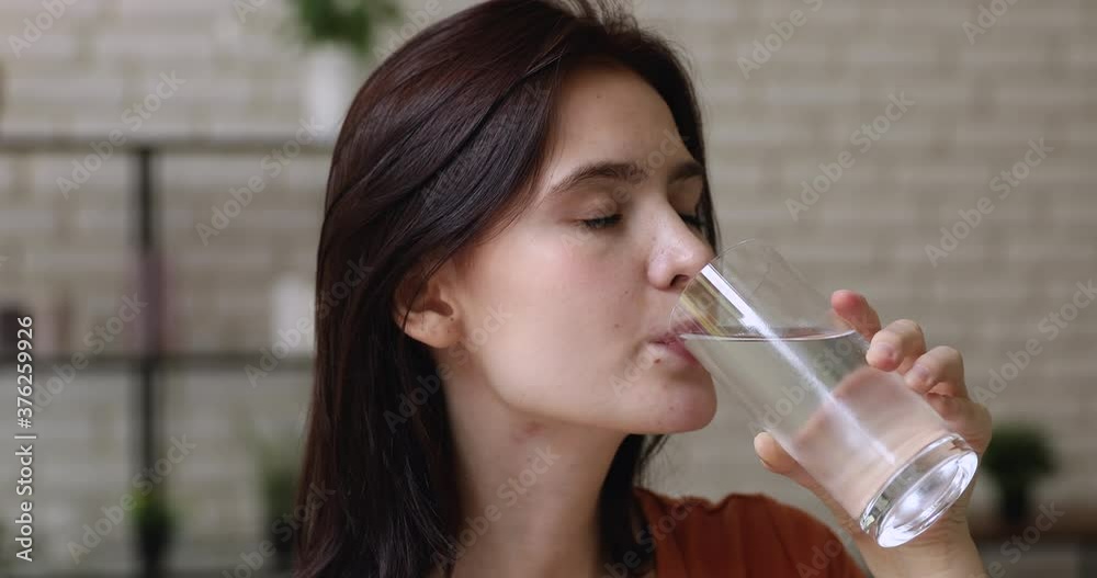 Close up young 20s woman holding glass drinking water closed eyes reduces thirst take everyday vitamin pills makes sip standing alone indoor. Improve body balance, dieting, healthy lifestyle concept Close up young 20s woman holding glass drinking water closed eyes reduces thirst take everyday vitamin pills makes sip standing alone indoor. Improve body balance, dieting, healthy lifestyle concept