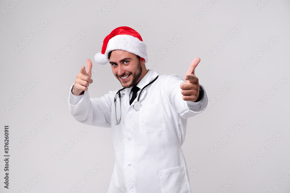 Young handsome doctor in white uniforme and Santa Claus hat standing in studio on white background smile and finger in camera Young handsome doctor in white uniforme and Santa Claus hat standing in studio on white background smile and finger in camera