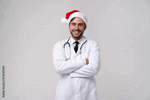 Young handsome doctor in white uniforme and Santa Claus hat standing in studio on white background loking at camera abd teeth smiling photo