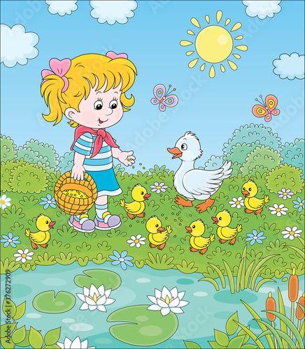 Fotografie, Obraz Little girl feeding a white duck and small yellow ducklings among flowers by a p