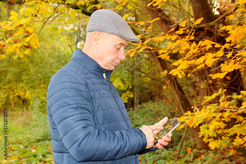 tinted profile photo of an elderly man in a gray cap and a blue jacket, who is standing in an autumn park among yellow and orange leaves of maple trees, is phoning