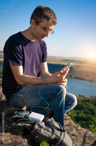 Man on a hike uses smartphone while charging from the power bank on the rock at dawn. Healthy lifestyle and communication.