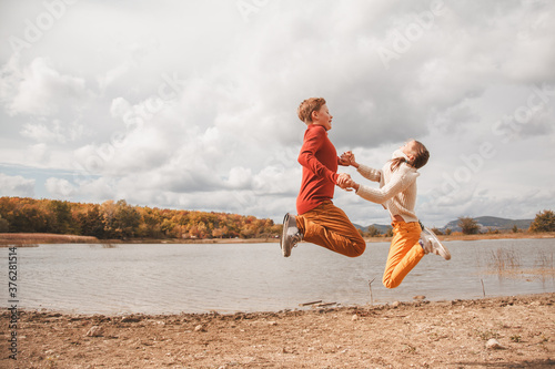 Preteen kids, boy and girl jumping in fall park with lake. Friends wearing warm knit sweaters