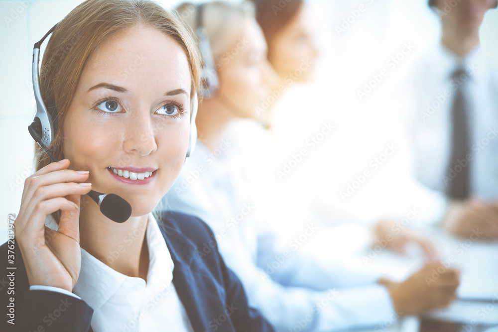 Cheerful smiling business woman with headphones consulting clients. Group of diverse phone operators at work in sunny office.Call center and business people concept Cheerful smiling business woman with headphones consulting clients. Group of diverse phone operators at work in sunny office.Call center and business people concept