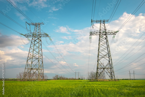 High voltage lines and power pylons in a flat and green agricultural landscape on a sunny day with clouds in the blue sky. Cloudy and rainy. Wheat is growing