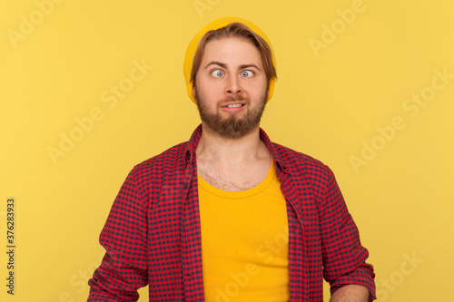 Funny dumb hipster bearded guy in beanie hat and checkered shirt making silly face with eyes crossed, looking at camera stupid brainless expression. indoor studio shot isolated on yellow background