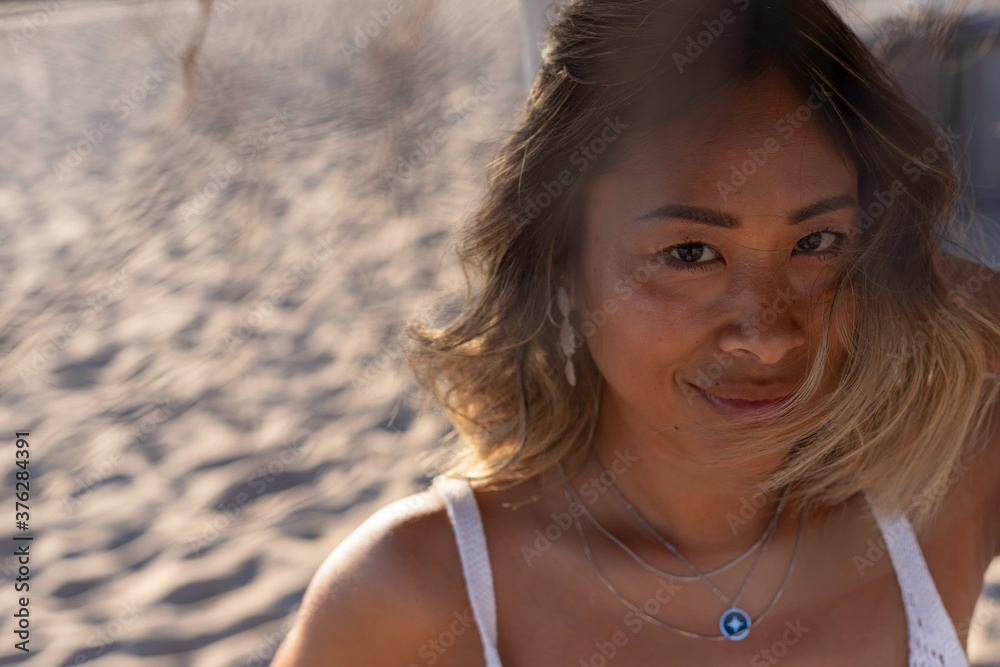 Close up portrait of a young beautiful Thai woman wearing a wooden hat and stylish necklace relaxing alone on the beach near the Atlantic Ocean. Woman's freedom lifestyle concept. Lisbon, Portugal