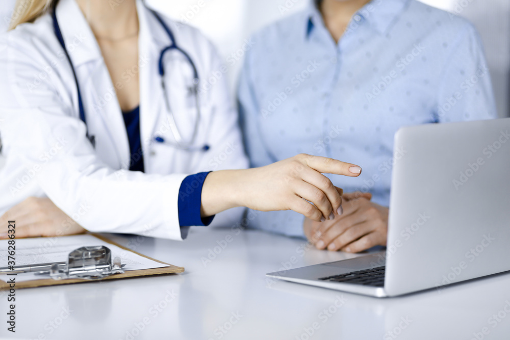 Unknown woman- doctor and her patient are discussing patient's blood test, while sitting together at the desk in the cabinet in a clinic. Female physician, with a stethoscope, is using a laptop Unknown woman- doctor and her patient are discussing patient's blood test, while sitting together at the desk in the cabinet in a clinic. Female physician, with a stethoscope, is using a laptop