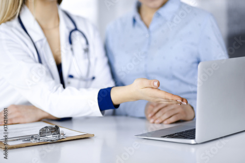 Unknown woman- doctor and her patient are discussing patient's blood test, while sitting together at the desk in the cabinet in a clinic. Female physician, with a stethoscope, is using a laptop