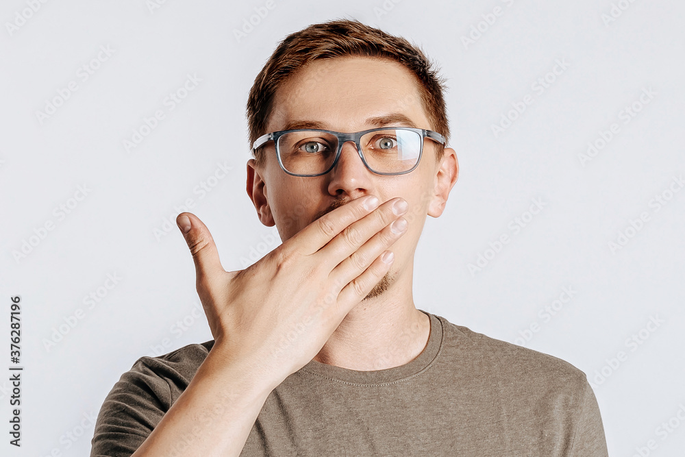 Young handsome man with glasses a brunette beard posing on isolated gray background. Place for advertising. The surprised man is shocked and covers his mouth with his hand. Bad surprise news. Young handsome man with glasses a brunette beard posing on isolated gray background. Place for advertising. The surprised man is shocked and covers his mouth with his hand. Bad surprise news.