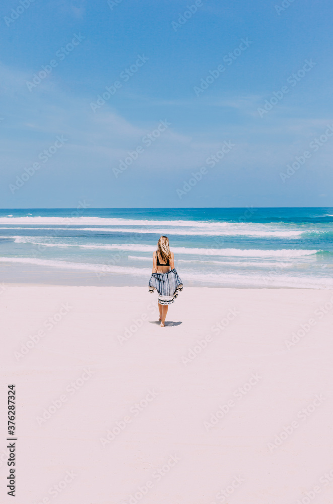 Young woman goes to the sea in beach clothes. Bright blue sky. Summertime, tropical island. 
