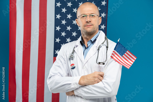 Confident middle aged doctor in eyeglasses and whitecoat holding small US flag