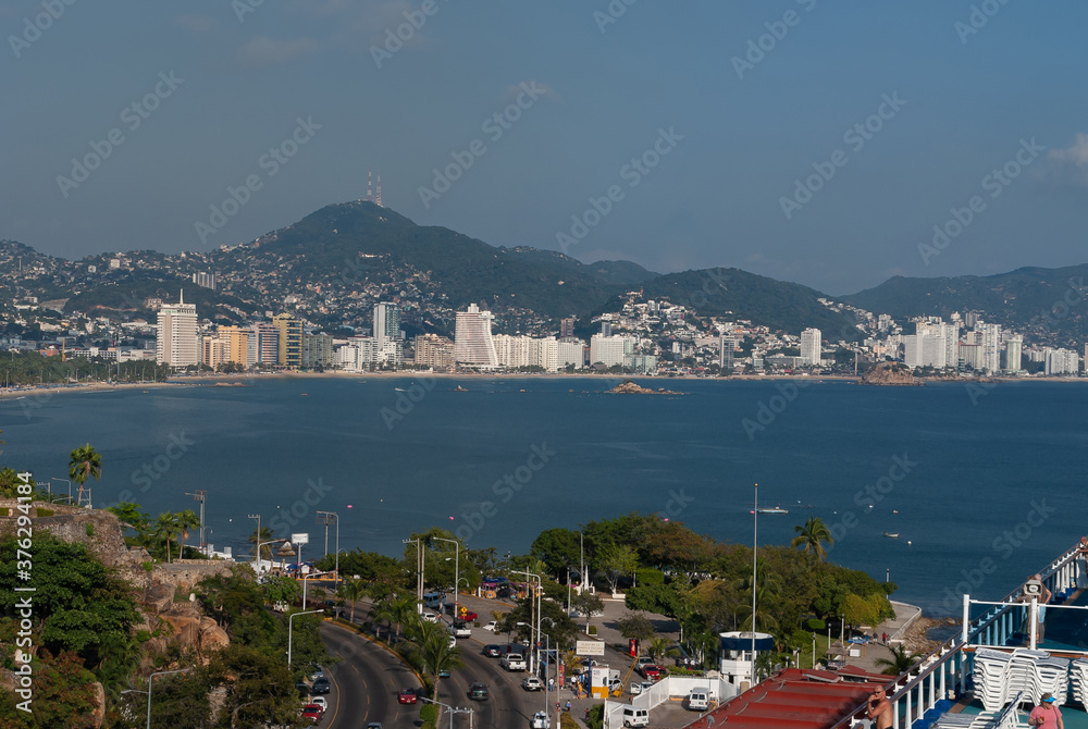 Acapulco, Mexico - November 25, 2008: The blue water bay with beach and highrise buildings along shoreline. Backdrop of green hills loaded with houses under blue sky. Front is freeway. 