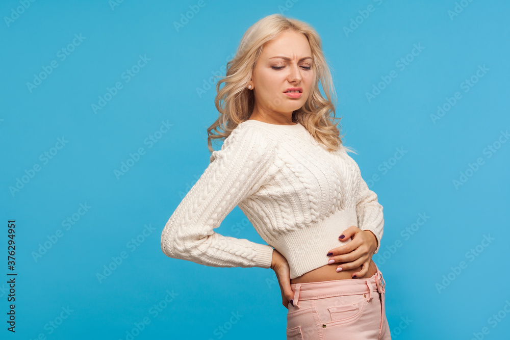 Depressed unhappy blond woman massaging numb back, suffering from kidney pain, spine inflammation. Indoor studio shot isolated on blue background Depressed unhappy blond woman massaging numb back, suffering from kidney pain, spine inflammation. Indoor studio shot isolated on blue background