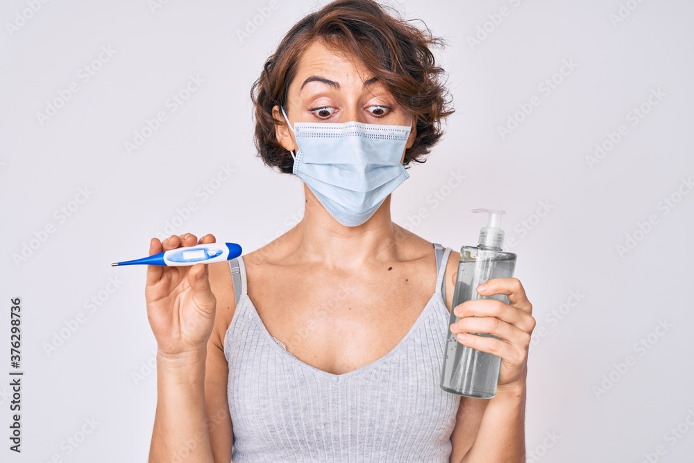 Young hispanic woman wearing medical mask, hand sanitizer gel and thermometer making fish face with mouth and squinting eyes, crazy and comical.