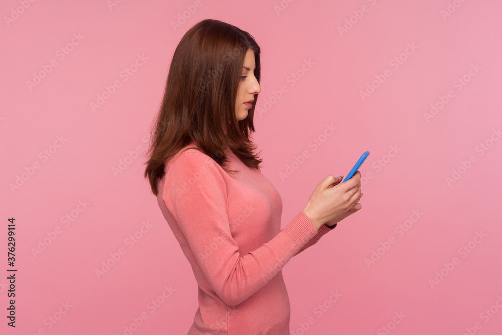 Side view serious concentrated brunette woman holding cell phone in hands, chatting or checking e-mail, gadget addiction. Indoor studio shot isolated on pink background Side view serious concentrated brunette woman holding cell phone in hands, chatting or checking e-mail, gadget addiction. Indoor studio shot isolated on pink background