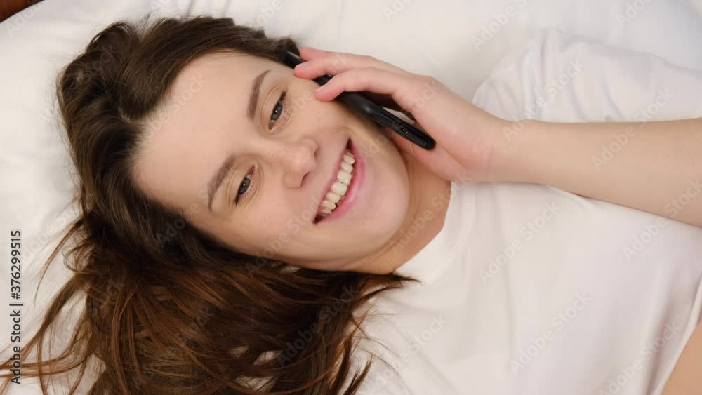Top view close up of smiling millennial woman talking mobile phone lying on comfortable white bed at home. Attractive young female having pleasant conversation chatting by mobile with friend Top view close up of smiling millennial woman talking mobile phone lying on comfortable white bed at home. Attractive young female having pleasant conversation chatting by mobile with friend