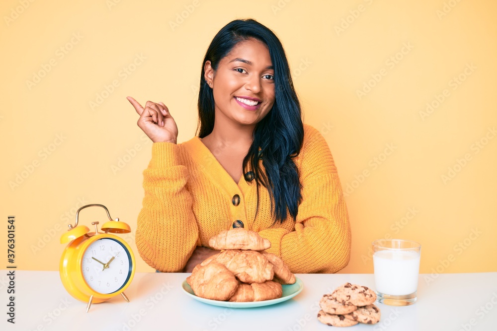 Beautiful latin young woman with long hair sitting on the table having breakfast smiling happy pointing with hand and finger to the side Beautiful latin young woman with long hair sitting on the table having breakfast smiling happy pointing with hand and finger to the side