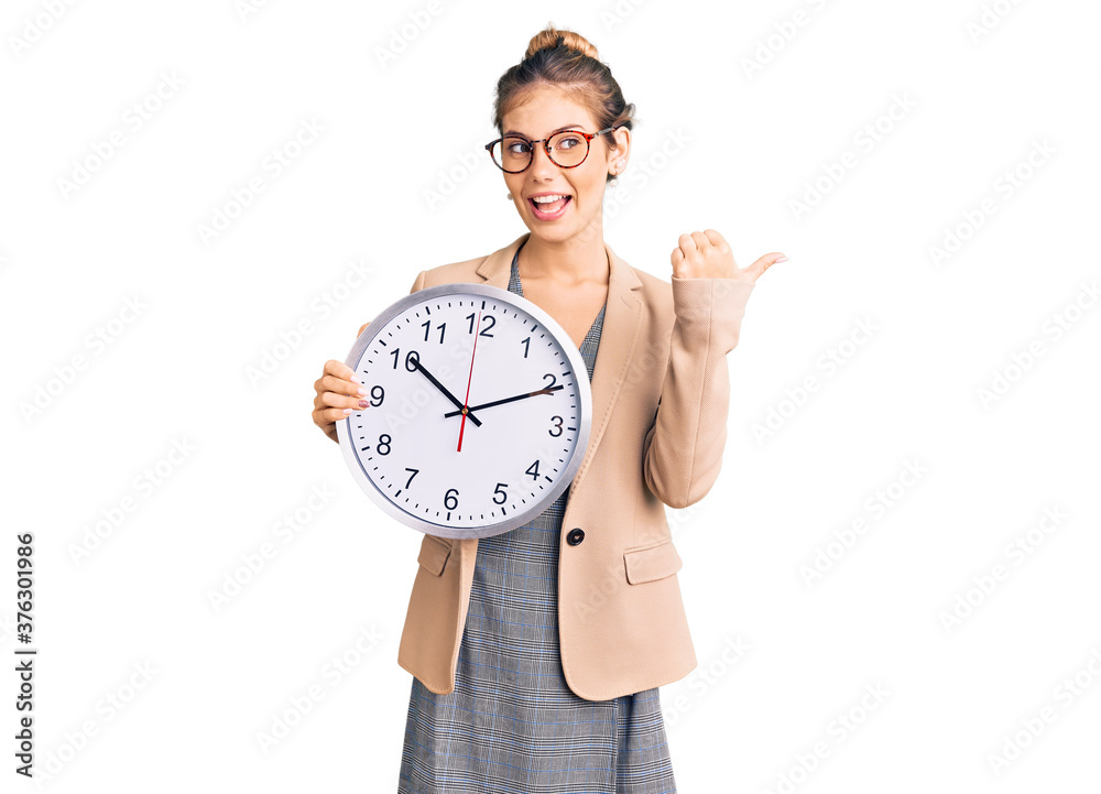 Beautiful caucasian woman with blonde hair wearing business jacket and holding clock pointing thumb up to the side smiling happy with open mouth Beautiful caucasian woman with blonde hair wearing business jacket and holding clock pointing thumb up to the side smiling happy with open mouth