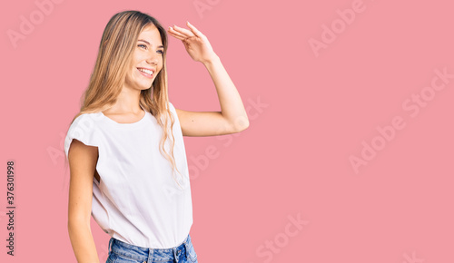 Beautiful caucasian woman with blonde hair wearing casual white tshirt very happy and smiling looking far away with hand over head. searching concept.