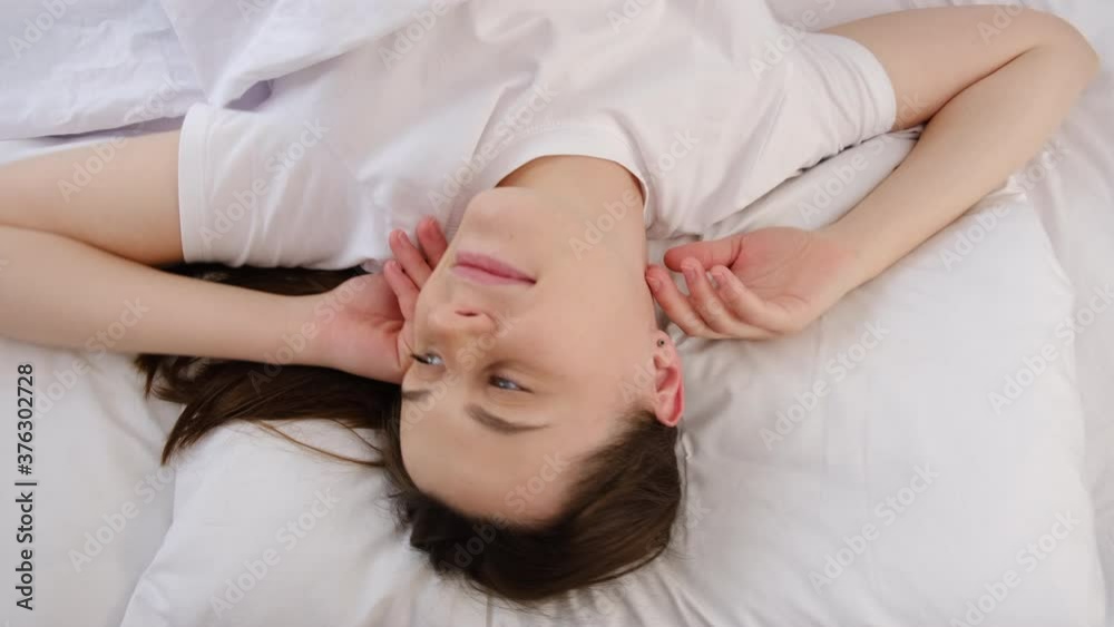 Top view portrait of Caucasian young pretty woman smiling while lying in bed early in morning after waking up. Cute beautiful girl looking happy in bedroom on white pillow in bedroom at home Top view portrait of Caucasian young pretty woman smiling while lying in bed early in morning after waking up. Cute beautiful girl looking happy in bedroom on white pillow in bedroom at home
