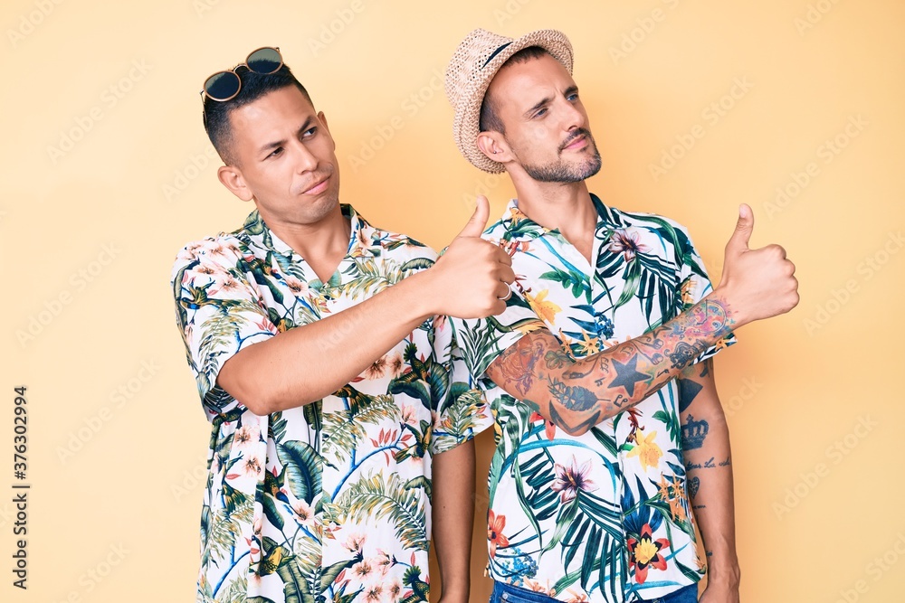 Young gay couple of two men wearing summer hat and hawaiian shirt looking proud, smiling doing thumbs up gesture to the side