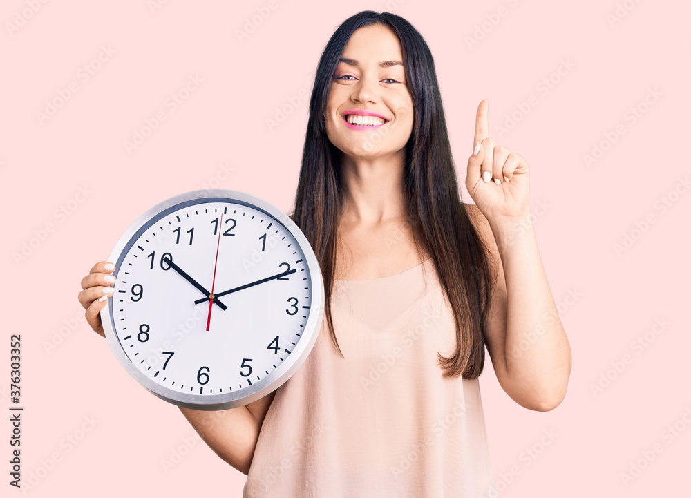 Young beautiful caucasian woman holding big clock surprised with an idea or question pointing finger with happy face, number one Young beautiful caucasian woman holding big clock surprised with an idea or question pointing finger with happy face, number one