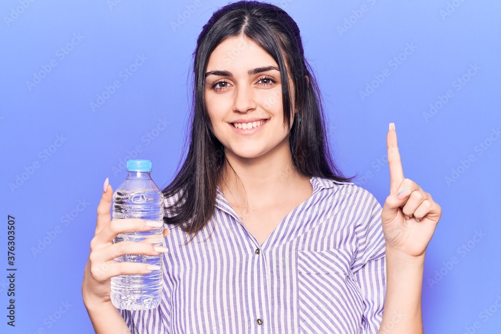 Young beautiful girl holding bottle of water smiling with an idea or question pointing finger with happy face, number one Young beautiful girl holding bottle of water smiling with an idea or question pointing finger with happy face, number one