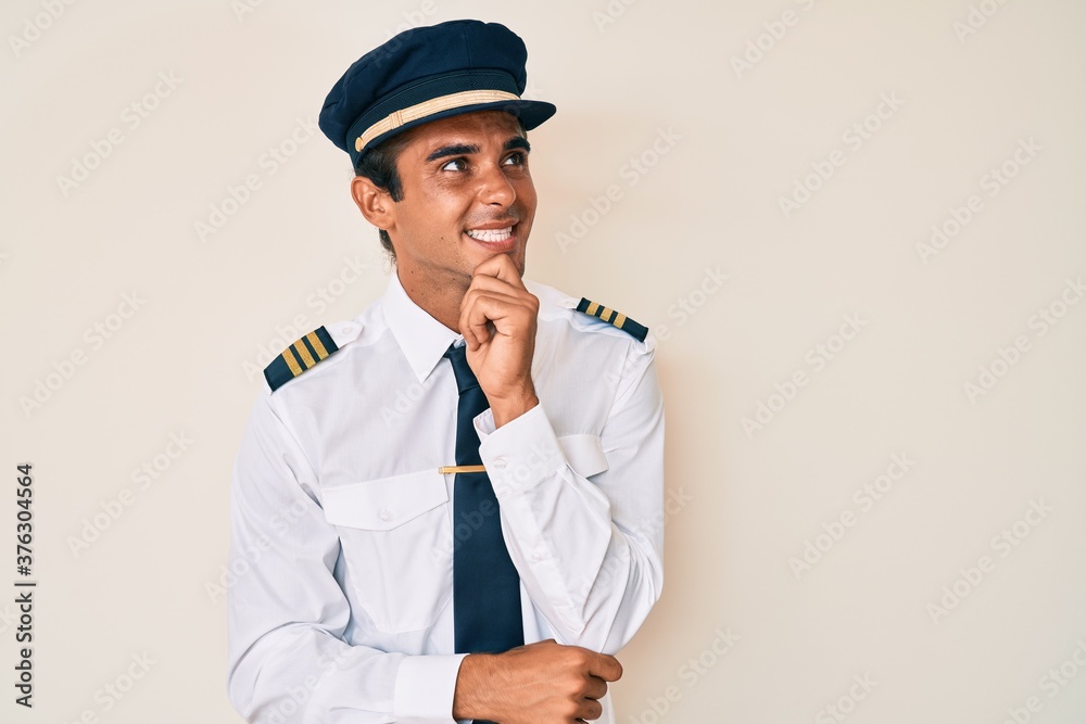 Young hispanic man wearing airplane pilot uniform with hand on chin thinking about question, pensive expression. smiling and thoughtful face. doubt concept. Young hispanic man wearing airplane pilot uniform with hand on chin thinking about question, pensive expression. smiling and thoughtful face. doubt concept.