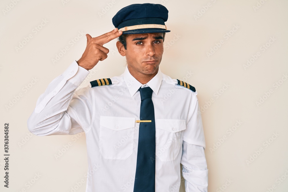 Young hispanic man wearing airplane pilot uniform shooting and killing oneself pointing hand and fingers to head like gun, suicide gesture.