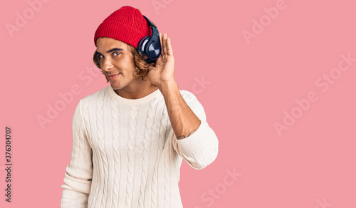 Young hispanic man listening to music using headphones smiling with hand over ear listening an hearing to rumor or gossip. deafness concept.