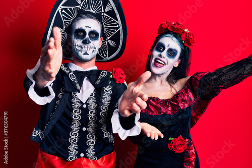 Young couple wearing mexican day of the dead costume over red looking at the camera smiling with open arms for hug. cheerful expression embracing happiness.