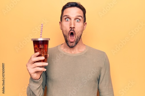 Young handsome man drinking cola refreshment beverage over isolated yellow background scared and amazed with open mouth for surprise, disbelief face photo