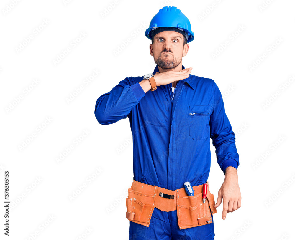 Young handsome man wearing worker uniform and hardhat in hurry pointing to watch time, impatience, looking at the camera with relaxed expression