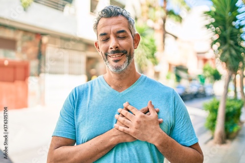 Middle age grey-haired man wearing casual clothes at street of city smiling with hands on chest with closed eyes and grateful gesture on face. health concept.