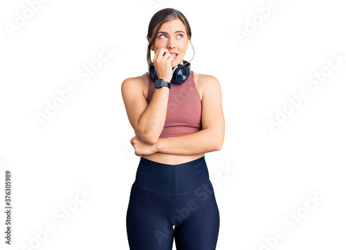 Beautiful caucasian young woman wearing gym clothes and using headphones looking stressed and nervous with hands on mouth biting nails. anxiety problem.