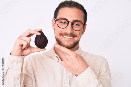 Young handsome man holding avocado smiling happy pointing with hand and finger