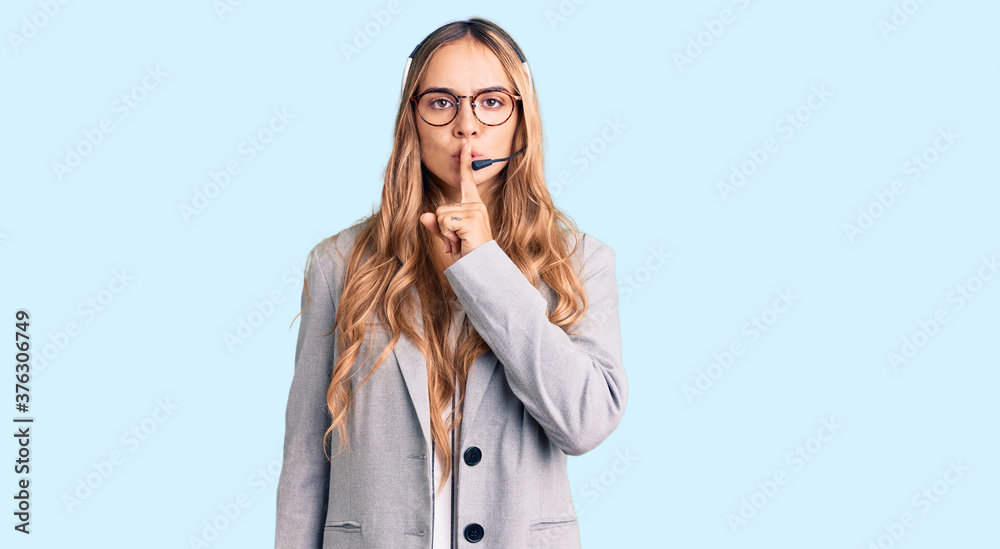 Young beautiful blonde woman wearing call center agent headset asking to be quiet with finger on lips. silence and secret concept.
