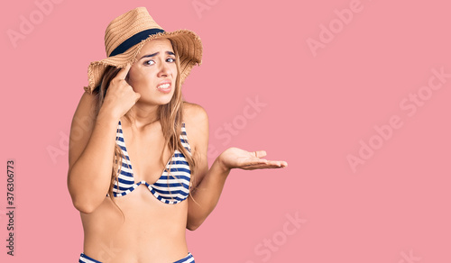 Young beautiful blonde woman wearing bikini and hat confused and annoyed with open palm showing copy space and pointing finger to forehead. think about it. photo