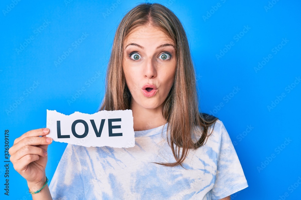 Young blonde girl holding paper with love message scared and amazed with open mouth for surprise, disbelief face Young blonde girl holding paper with love message scared and amazed with open mouth for surprise, disbelief face