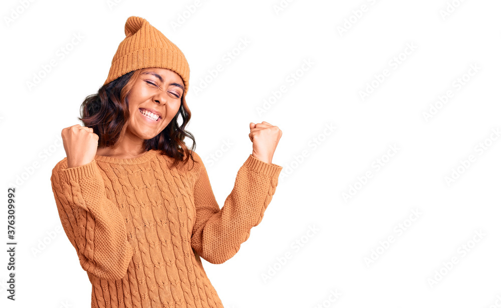 Young beautiful mixed race woman wearing wool sweater and winter hat very happy and excited doing winner gesture with arms raised, smiling and screaming for success. celebration concept. Young beautiful mixed race woman wearing wool sweater and winter hat very happy and excited doing winner gesture with arms raised, smiling and screaming for success. celebration concept.