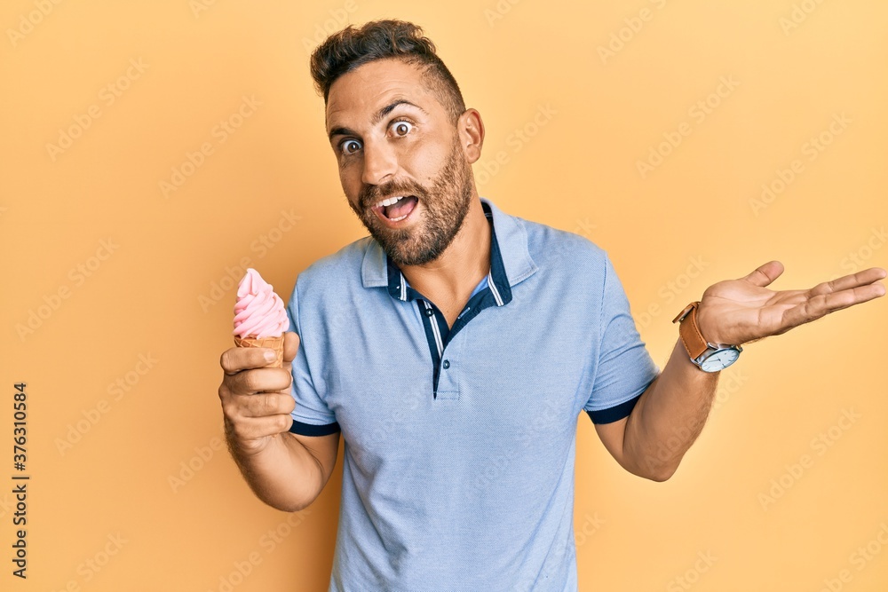 Handsome man with beard eating ice cream cone celebrating achievement with happy smile and winner expression with raised hand Handsome man with beard eating ice cream cone celebrating achievement with happy smile and winner expression with raised hand