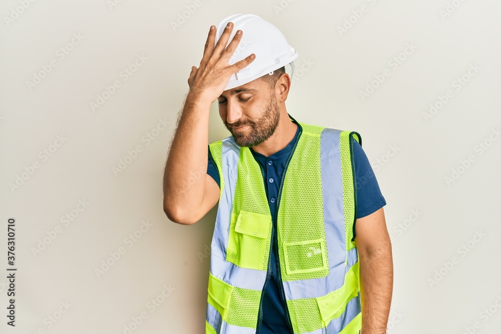 Handsome man with beard wearing safety helmet and reflective jacket surprised with hand on head for mistake, remember error. forgot, bad memory concept.