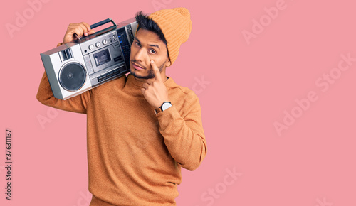 Handsome latin american young man holding boombox, listening to music pointing to the eye watching you gesture, suspicious expression