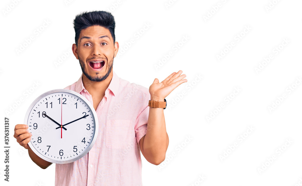 Handsome latin american young man holding big clock celebrating victory with happy smile and winner expression with raised hands Handsome latin american young man holding big clock celebrating victory with happy smile and winner expression with raised hands