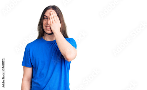 Young adult man with long hair wearing swimwear covering one eye with hand, confident smile on face and surprise emotion.