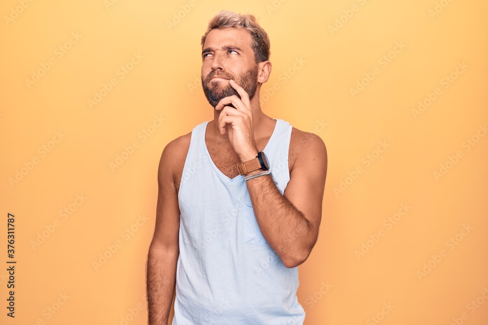 Young handsome blond man wearing casual sleeveless t-shirt over isolated yellow background thinking concentrated about doubt with finger on chin and looking up wondering Young handsome blond man wearing casual sleeveless t-shirt over isolated yellow background thinking concentrated about doubt with finger on chin and looking up wondering