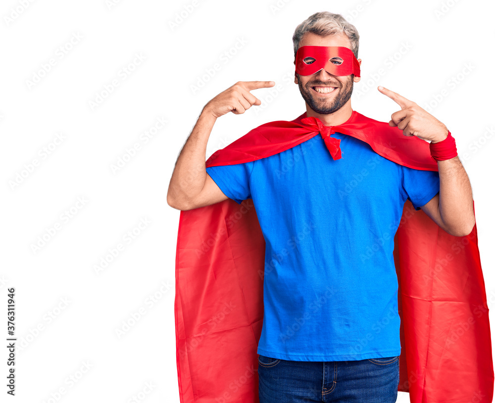 Young blond man wearing super hero custome smiling cheerful showing and pointing with fingers teeth and mouth. dental health concept. Young blond man wearing super hero custome smiling cheerful showing and pointing with fingers teeth and mouth. dental health concept.