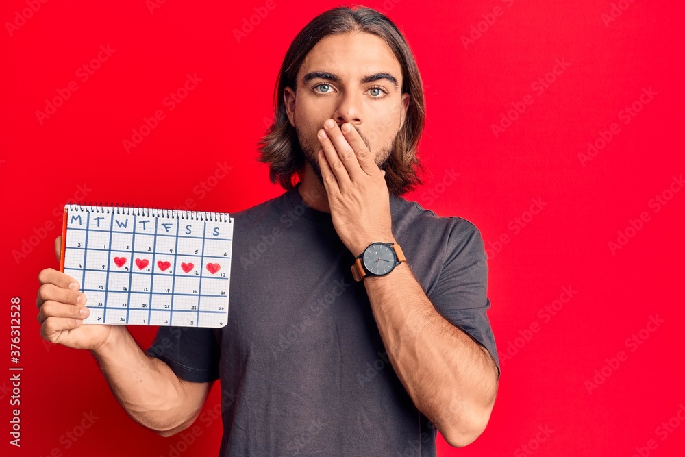Young handsome man holding heart calendar covering mouth with hand, shocked and afraid for mistake. surprised expression Young handsome man holding heart calendar covering mouth with hand, shocked and afraid for mistake. surprised expression