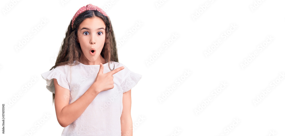 Cute hispanic child girl wearing casual white tshirt surprised pointing with finger to the side, open mouth amazed expression.